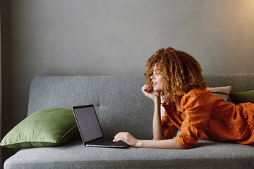Young woman with curly hair in orange blouse working on laptop at home with green pillows, relaxed atmosphere, focus on productivity and comfort