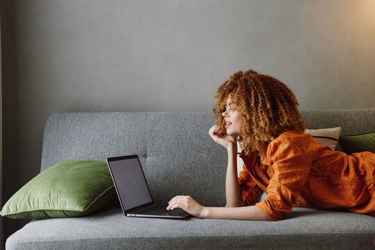 Young woman with curly hair in orange blouse working on laptop at home with green pillows, relaxed atmosphere, focus on productivity and comfort