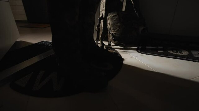 Military soldier returns home after service or war. Close-up legs of military man in tactical boots who enters an apartment, takes off military tactical backpack and places it on floor. Armed forces.