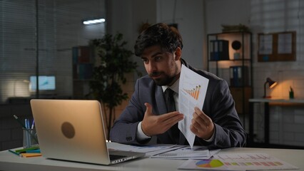 Adult business man manager sitting at desk working on laptop in office overtime in evening, talks online by video call, shows papers with infographic.