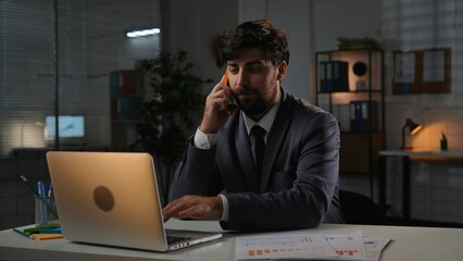 Adult business man manager sitting at desk working on laptop in office overtime in evening, typing on computer answers call, talking on smartphone.