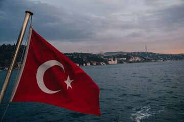 Turkish flag waving in the wind with Istanbul cityscape and Bosphorus in background, from a tour boat sailing through the strait during a cloudy sunset, Turkey