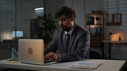 Adult business man manager sitting at desk working on laptop in office overtime in evening, typing on computer doing research, reading information.