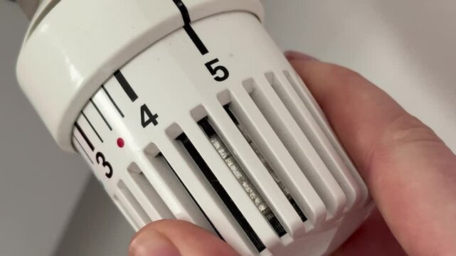 A person adjusting the white thermostat on a white radiator at home to raise the room temperature during the cold season.
