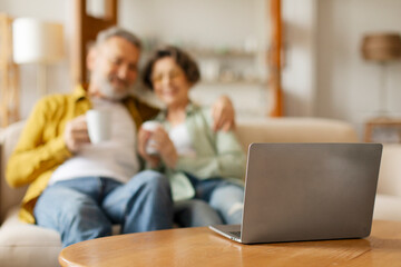 Focus on computer while senior couple watching tv or film online on laptop and drinking tea, resting on comfortable sofa