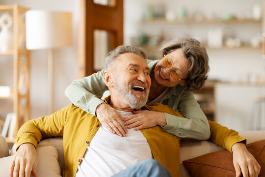 Loving romantic senior couple enjoying their relationships and long lasting marriage, woman embracing her husband while man sitting on sofa