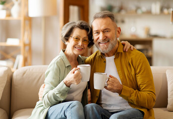 Happy senior couple drinking coffee together, enjoying cozy moments over cup of hot drink and smiling at camera, sitting on couch at home