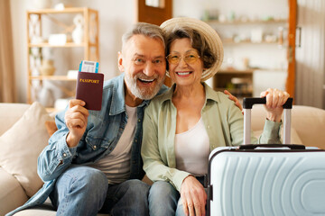 New trip, finally. Excited senior couple holding travel tickets and passports, sitting with packed suitcases on sofa and smiling at camera