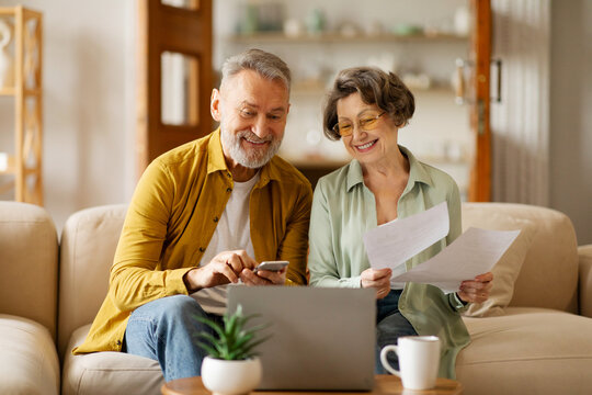 Positive senior spouses checking financial documents and calculating family budget, using laptop and cellphone, sitting at table in living room