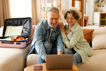 Happy senior couple planning vacation travel with laptop, booking tickets or hotel accommodation on web, sitting with unpacked suitcase at home