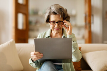 Positive senior woman using laptop and touching glasses, surfing internet or video calling with...