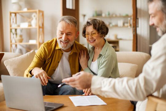 Real estate agent showing house plans on laptop to senior couple clients, sitting on sofa indoors. Agency service offer concept
