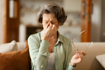 Unhappy grey haired senior woman massaging nose bridge, suffering from eyestrain, taking off...