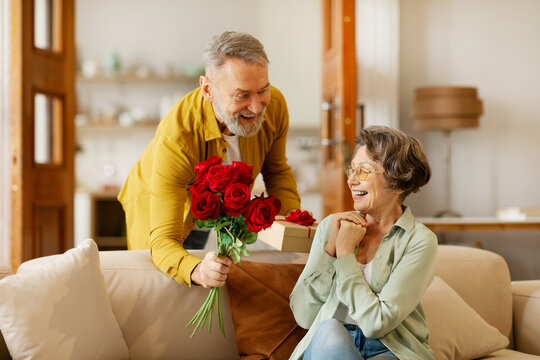 Loving senior man giving bouquet of flowers and gift box to surprised wife, sitting on sofa at home. Birthday, anniversary or Valentine's day celebration