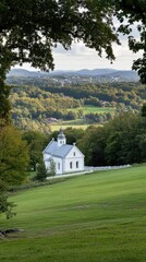 Obraz premium A quaint white and gray church with a black roof sits in a green field, surrounded by trees displaying autumn colors under a cloudy sky