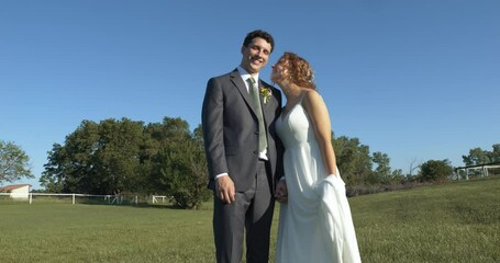 A joyful bride and groom kissing in a sunlit field, under tree shade, sharing smiles and laughter, as they celebrate their special day in a picturesque outdoor setting surrounded by nature.