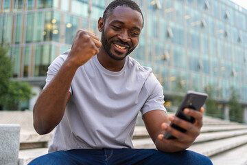 Happy African American man euphoric winner with smartphone on street in city. Person guy looking at...