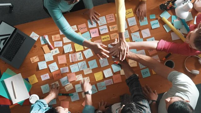 Top down view of designer standing at meeting with sticky note and putting hand together. Aerial view of team make stack of hand to celebrate successful project or after brainstorming idea. Symposium.
