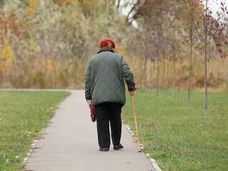 A lonely elderly woman with red hair walks with a stick along the alley of an autumn park. Breathe fresh air in nature. Active aging. Healthy lifestyle in old age