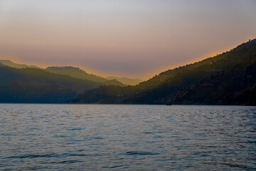 The evening blue waters of the lake create a serene atmosphere, complemented by multiple layers of mountains. Evening yellow-orange light shines on the mountain tops, adding warmth to Tehri Lake.