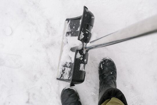 An unrecognizable person in snow boots holding a plastic shovel in order to clean, remove snow from a sidewalk for safety