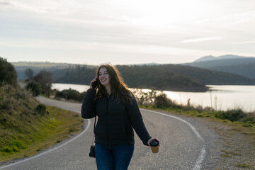 Young woman walking on a country road, talking on smartphone and holding coffee