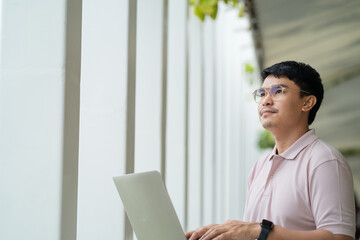 A man wearing glasses is typing on a laptop while looking out the window. He is focused on his work, but also seems to be enjoying the view outside