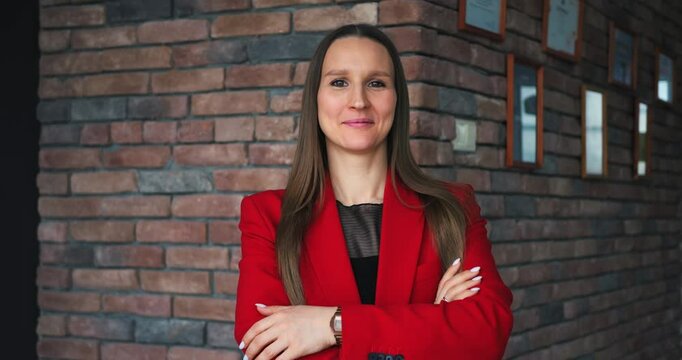 Confident businesswoman in red blazer stands with arms crossed, projecting success in brick backdrop, empowering businesswoman, fueling businesswoman motivation for global reach and innovation.