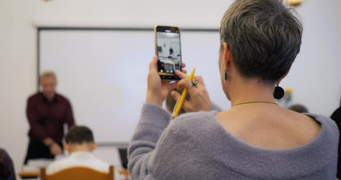 Mature woman capturing classroom moment on smartphone during educational session, blending modern technology with traditional learning environment. Attendee recording lecture using smartphone