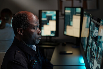 Fototapeta premium Mature African American security guard concentrating on watching cctv video on computer screen in surveillance room