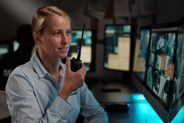 Young blond female officer of surveillance room reporting situation on walkie-talkie while looking...