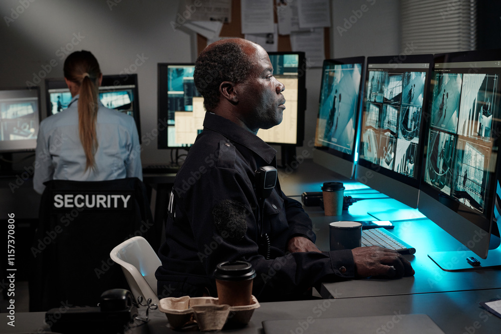 Poster side view of serious african american officer of surveillance room looking at computer screen with c