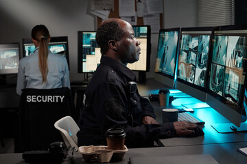 Side view of serious African American officer of surveillance room looking at computer screen with...