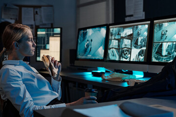 Young blond female officer with cup of coffee and sandwich watching cctv video on desktop computer screen at lunch break
