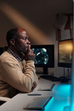 Pensive African American male programmer keeping fingers by mouth and looking at computer screen while trying to decode data