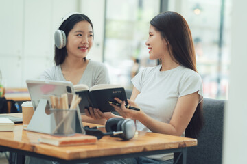 Fototapeta premium Smiling Friends: Two young women, headphones on, share a lighthearted moment over a book while studying in a modern coffee shop. 