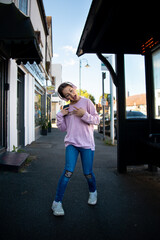 Young girl posing playfully on a vibrant city street during the day.  A girl in a pink sweater joyfully poses with excitement on a lively urban sidewalk, surrounded by charming shops and a clear blue 