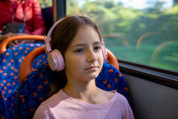 A girl with headphones sits quietly on a bus, immersed in her music while the lush green scenery passes by outside the window. The atmosphere is serene.
