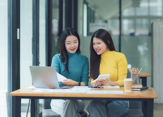 Collaborative Success: Two smiling young Asian businesswomen review documents and data on a laptop and tablet during a productive meeting in a modern office setting.  