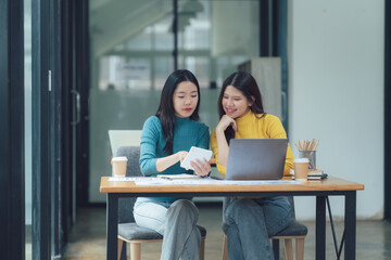 Collaborative Brainstorming: Two young female professionals work together in a modern office setting, collaborating on a project using a laptop and smartphone.