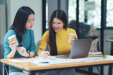 Collaborative Design Session: Two young Asian female designers brainstorm ideas, reviewing designs on a laptop and notepad during a productive meeting.  