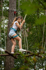 A young girl expertly balances on a swinging bridge high above the ground, surrounded by lush greenery, enjoying an adventurous day in a treetop park.
