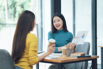 Collaborative Brainstorm: Two young businesswomen engage in a lively discussion over a cup of coffee, reviewing a chart and strategizing, showcasing the energy and dynamism of modern workplaces. 