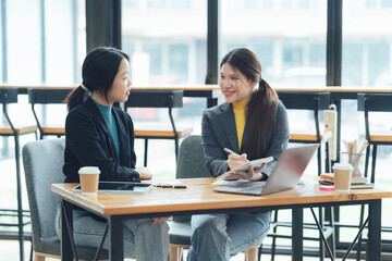 Businesswomen Meeting: Collaborative discussion between two Asian businesswomen in a modern office setting. Laptop, coffee cups, and notepad add to the professional atmosphere.  