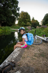 A cheerful girl wearing a helmet sits by a tranquil waterway, resting on the bank with her bike nearby.