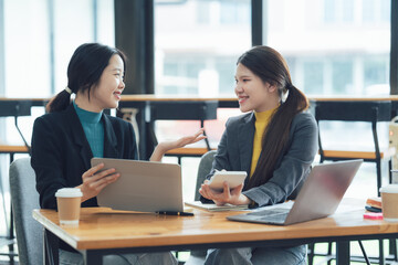 Dynamic Duo:  Two smiling Asian businesswomen collaborate on a project, engaging in lively discussion over laptops and documents in a modern office setting.  