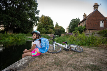 A girl in a helmet takes a break by a calm canal, enjoying nature. Her bicycle lies next to her as she gazes thoughtfully at the serene surroundings.