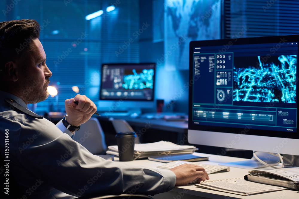 Poster young serious male operator in uniform looking at graphic data on computer screen while sitting by w