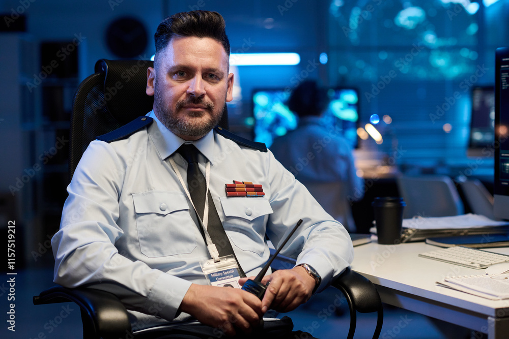 Poster confident mature officer wearing uniform and tie looking at camera while sitting by workplace with c