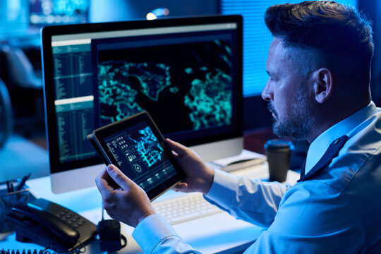 Serious mature man in uniform sitting by workplace in military command center and learning graphic data on tablet screen